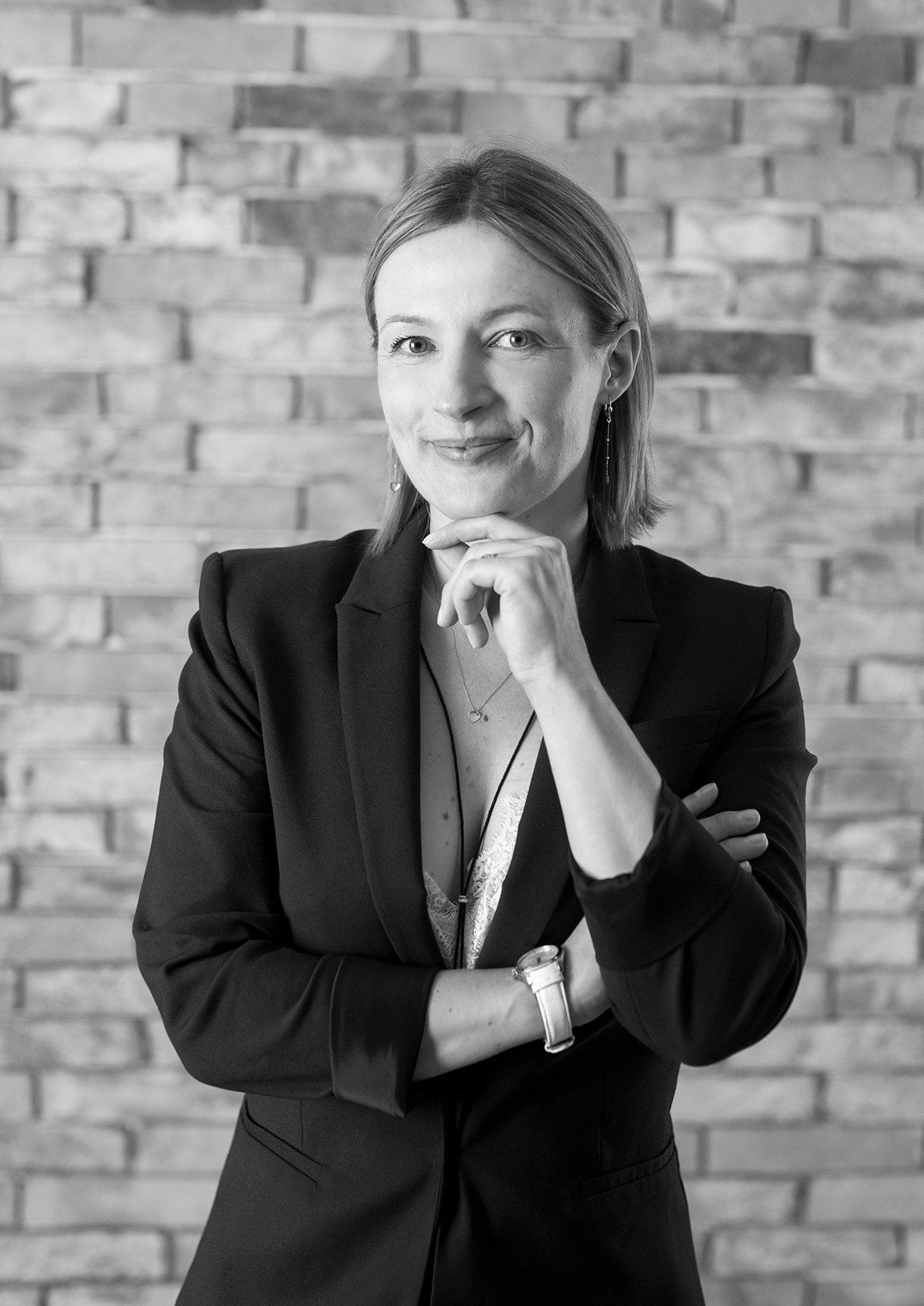 Czarno-białe portretowe ujęcie kobiety w czarnej marynarce, z ręką pod brodą i skrzyżowanymi ramionami, na tle ściany z cegieł