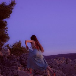 Grzegorz Grodzki Fotografia - Kobieta w zwiewnej sukience na skalistym wybrzeżu o zmierzchu, z widocznym drzewem po lewej stronie i górami w tle. Ujęcie z dołu, podkreślające dynamiczną pozę.