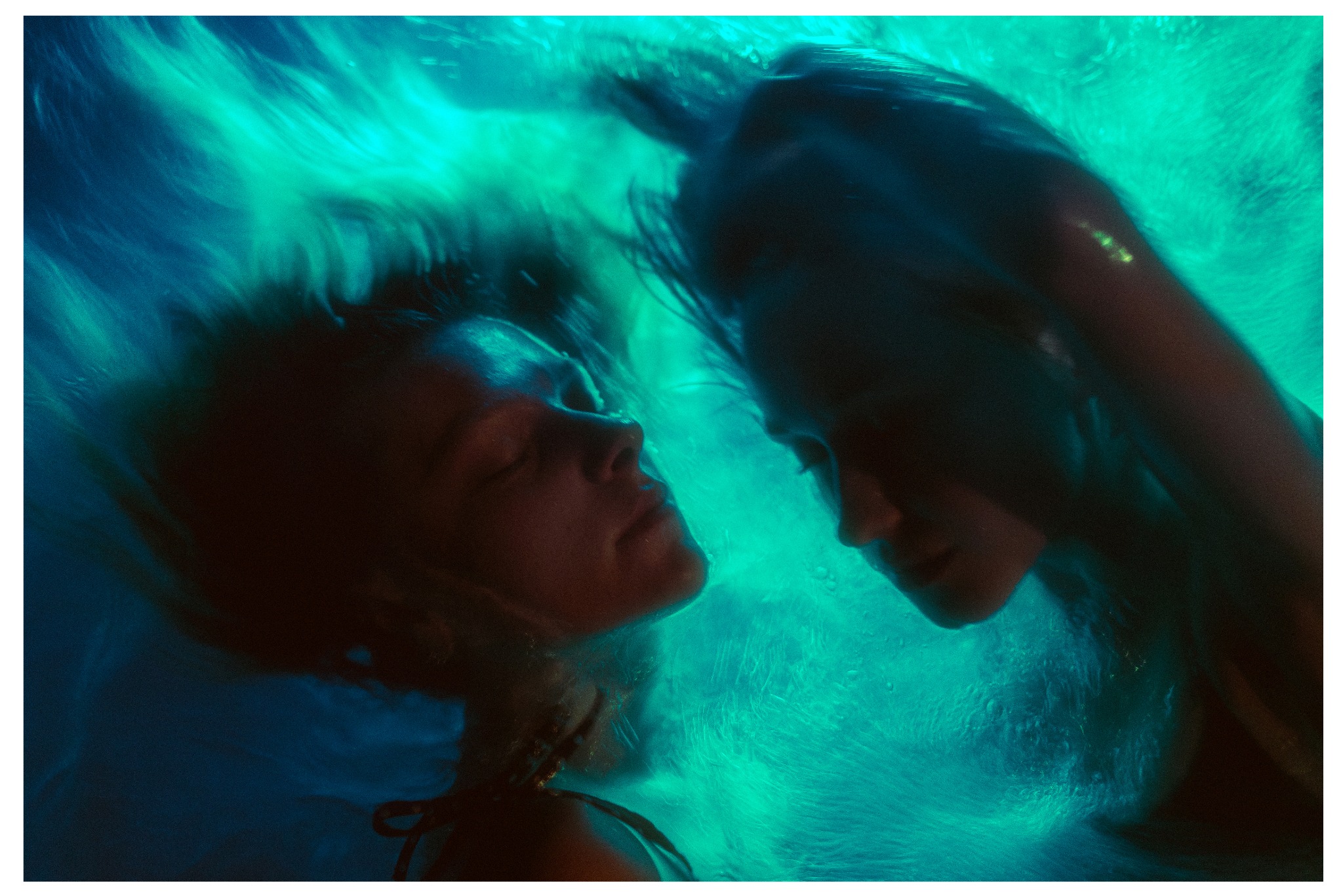 Artystyczne, podwodne ujęcie dwóch kobiet w turkusowej wodzie. Delikatne światło podkreśla kontury twarzy i włosów, tworząc eteryczny, niemal surrealistyczny efekt.