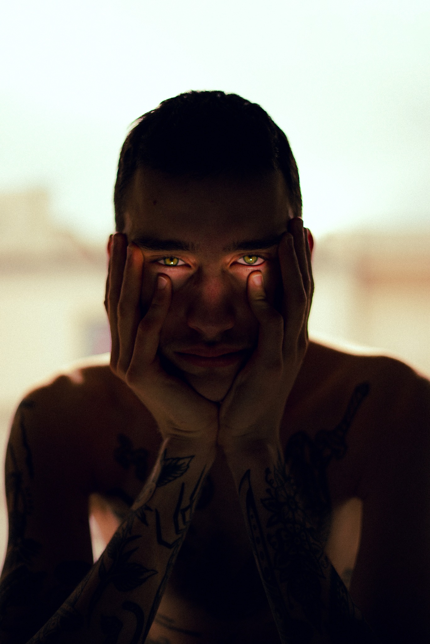 Mężczyzna z tatuażami na ramionach, opierający twarz na dłoniach, z intensywnym spojrzeniem zielonych oczu, w ciemnym otoczeniu z delikatnym, rozproszonym światłem.