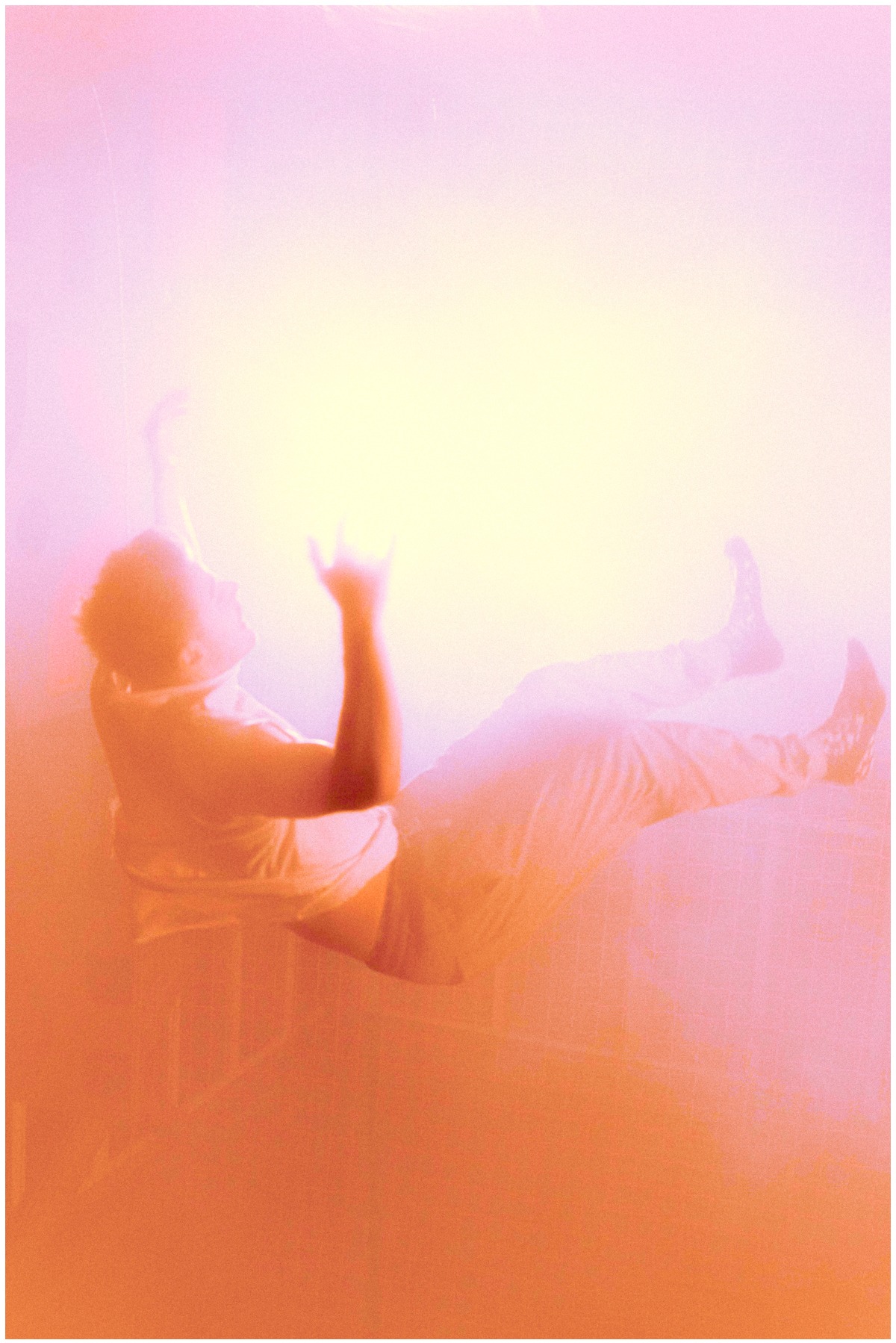 Artystyczne ujęcie mężczyzny w białym ubraniu, unoszącego ręce w górę w pomarańczowo-różowej, rozświetlonej przestrzeni. Nietypowe oświetlenie i kompozycja.