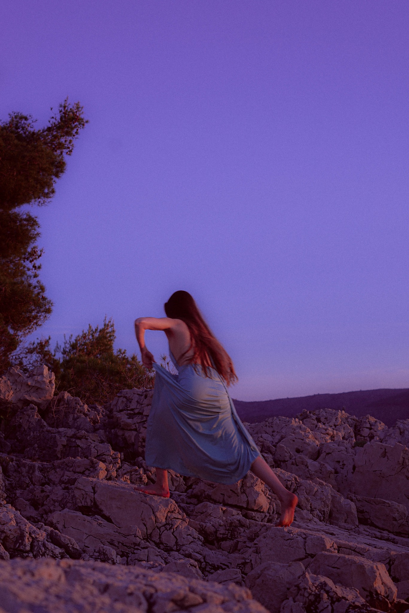 Kobieta w zwiewnej sukience na skalistym wybrzeżu o zmierzchu, z widocznym drzewem po lewej stronie i górami w tle. Ujęcie z dołu, podkreślające dynamiczną pozę.