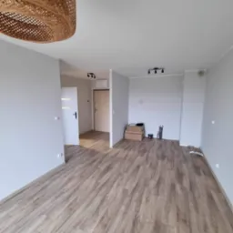 Wnętrze mieszkania po remoncie: świeżo pomalowane szare ściany, ułożona podłoga z paneli imitujących drewno, widoczna lampa z wikliny, drzwi wewnętrzne i wejściowe.