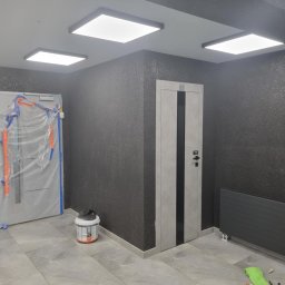 ARTDEK A.HASPARIAN B.BONDARCHUK A.KUCEWICZ SPÓŁKA CYWILNA - Wnętrze w trakcie prac: ściana pokryta ciemną, brokatową farbą, zabezpieczone folią drzwi wejściowe, zamontowane drzwi wewnętrzne z czarnym pasem, grzejnik panelowy i wiadro z farbą na podłodze...