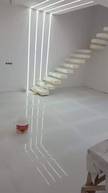 Białe wnętrze po szpachlowaniu i wylewce samopoziomującej, widoczne schody z zabezpieczonymi stopniami, wiadro farby i pionowe ledowe oświetlenie.