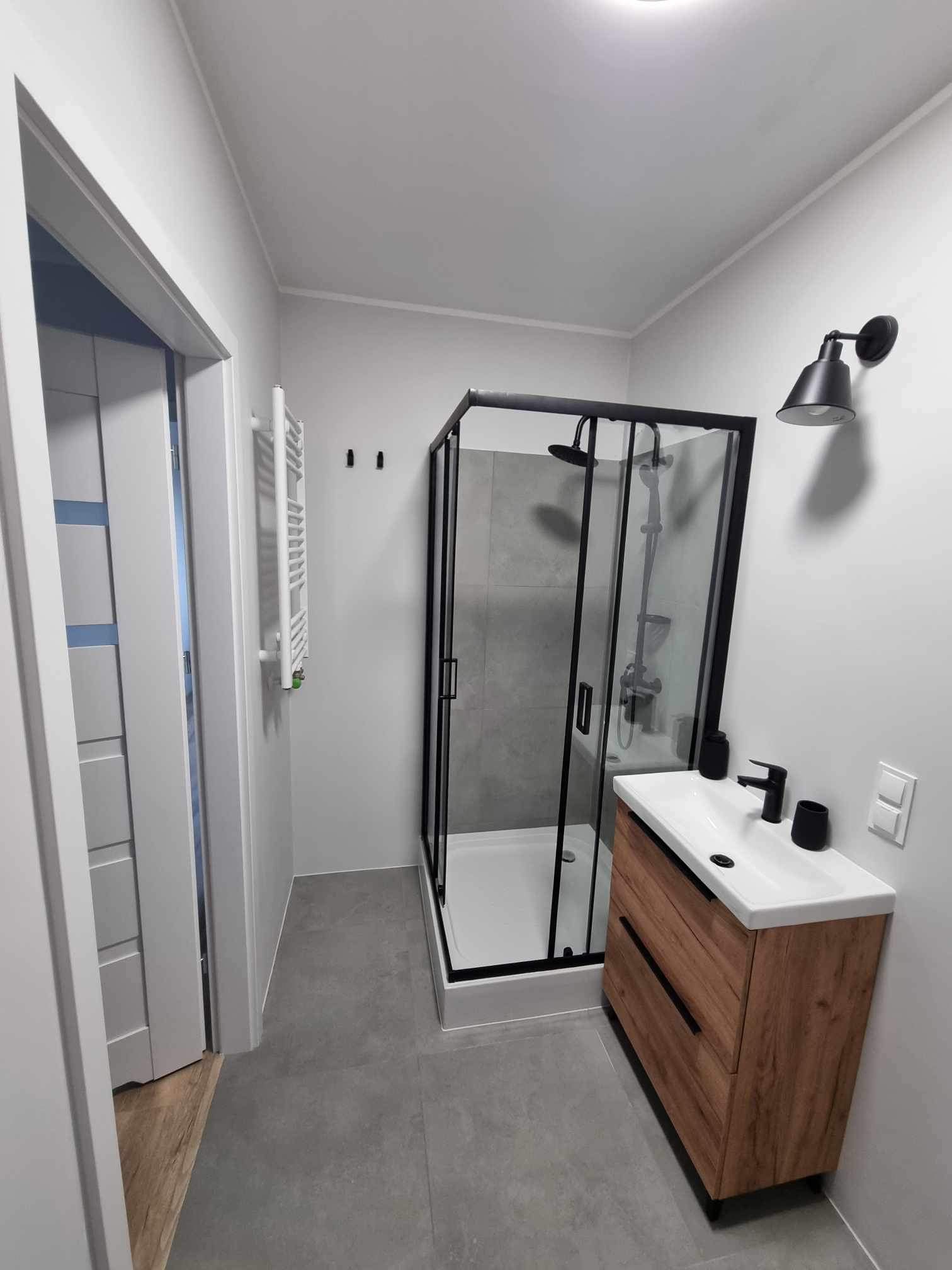 Nowoczesna łazienka z kabiną prysznicową w czarnej ramie, umywalką z szafką z drewna i czarną armaturą, szare płytki podłogowe i białe ściany z czarnymi haczykami i lampą.