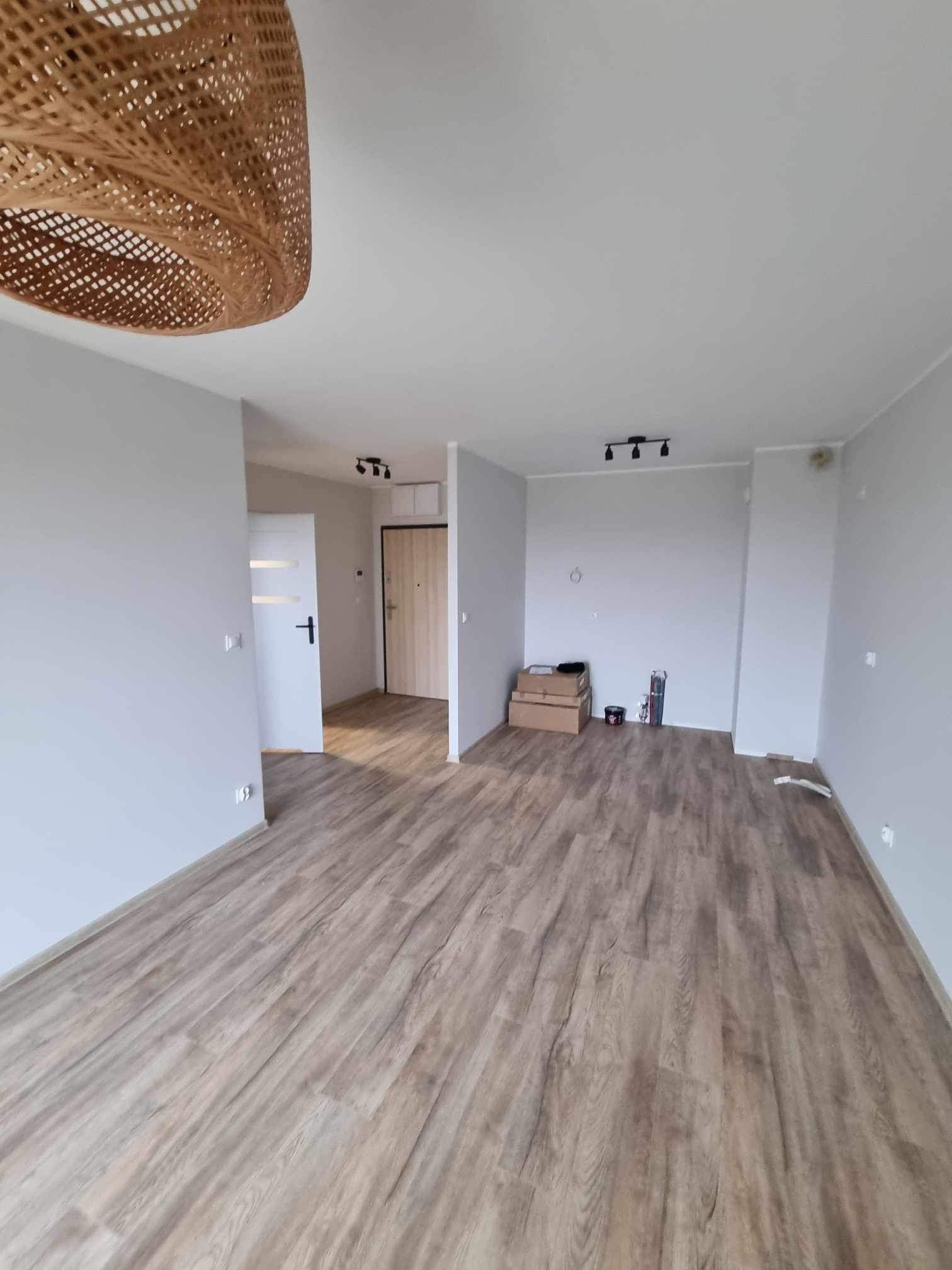 Wnętrze mieszkania po remoncie: świeżo pomalowane szare ściany, ułożona podłoga z paneli imitujących drewno, widoczna lampa z wikliny, drzwi wewnętrzne i wejściowe.