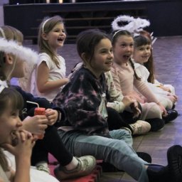 Iluzjonista Maciej Leksy - Grupa roześmianych dziewczynek w kostiumach aniołów siedzi rzędem na podłodze podczas wydarzenia. W tle dekoracje sceniczne i oświetlenie.