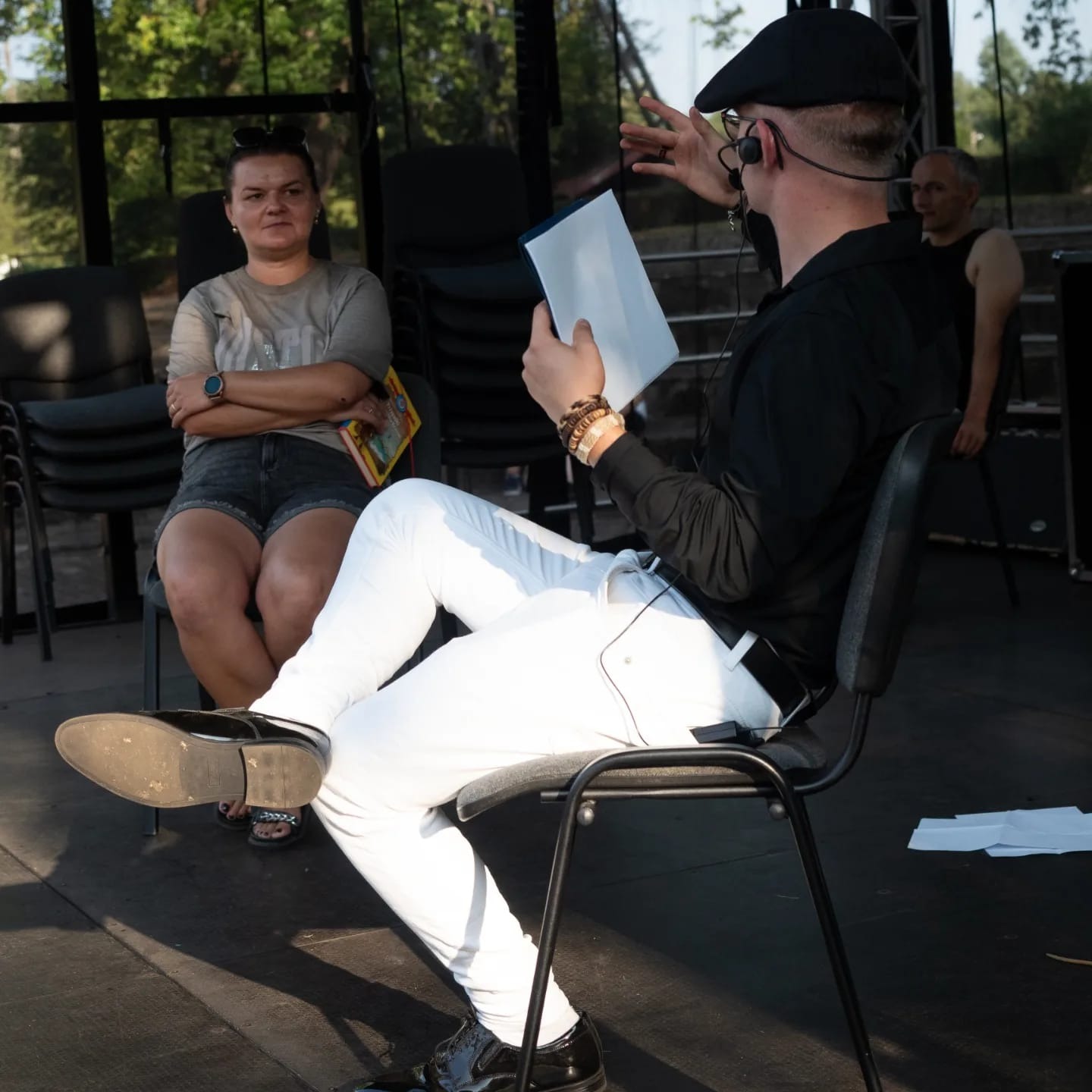 Mężczyzna w czarnej koszuli i białych spodniach, siedzący na krześle z mikrofonem, gestykuluje i trzyma kartkę papieru, w tle kobieta i mężczyzna.