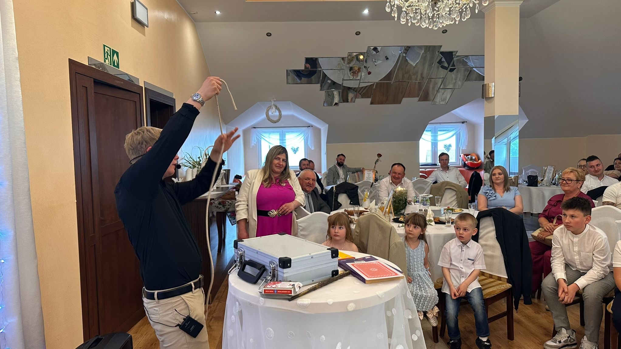 Iluzjonista w trakcie pokazu z liną dla publiczności na przyjęciu. Na stole rekwizyty, w tle obserwujący widzowie.