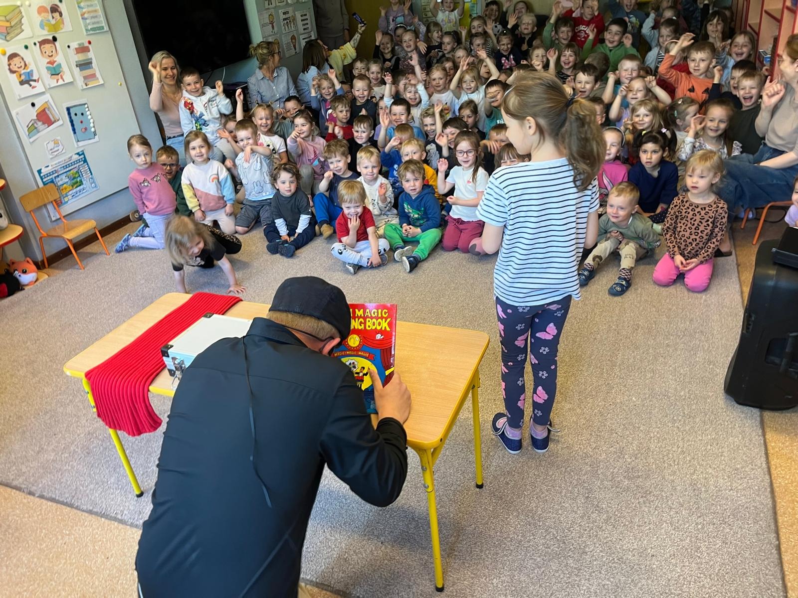 Iluzjonista prezentuje magiczną książkę przed grupą dzieci w przedszkolu, które z entuzjazmem reagują na pokaz. Stolik z rekwizytami.