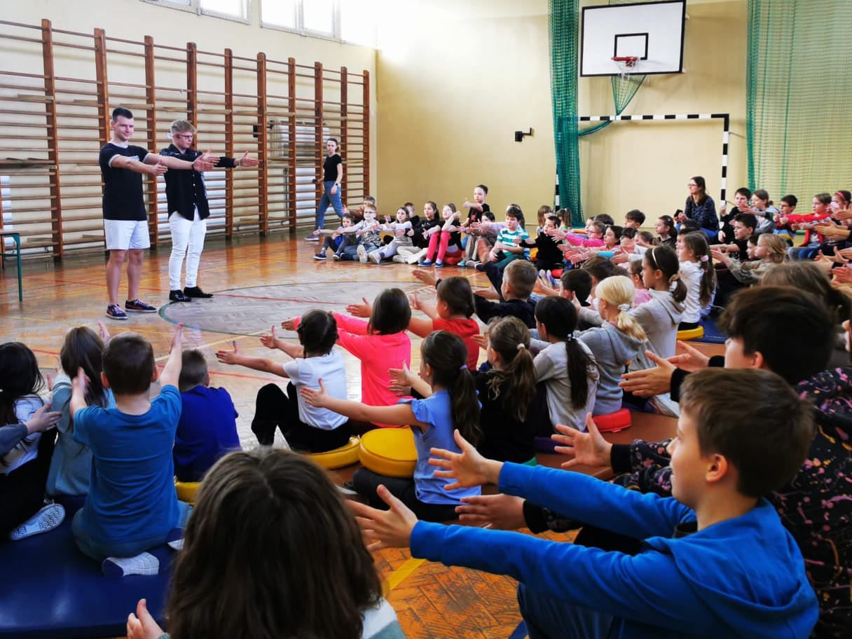 Dwóch mężczyzn prowadzi ćwiczenia rozciągające dla grupy dzieci siedzących na podłodze w sali gimnastycznej. W tle drabinki i kosz do koszykówki.