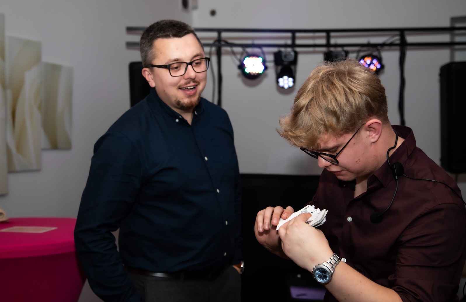 Iluzjonista w trakcie pokazu kart dla widza, oświetlenie sceniczne w tle. Mężczyzna w okularach z zaciekawieniem obserwuje trik.