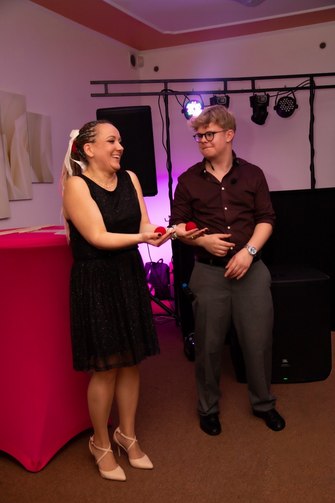 Uśmiechnięta iluzjonistka prezentuje lewitujące czerwone kulki, obok stoi asystent w okularach. W tle sprzęt nagłośnieniowy i oświetlenie sceniczne.