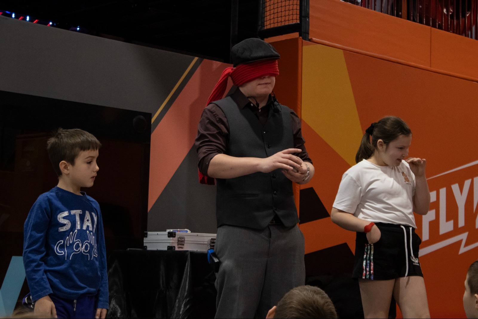 Iluzjonista z zasłoniętymi oczami prezentuje trik dwójce dzieci na tle pomarańczowej ściany. Na stole leży srebrna walizka z rekwizytami.