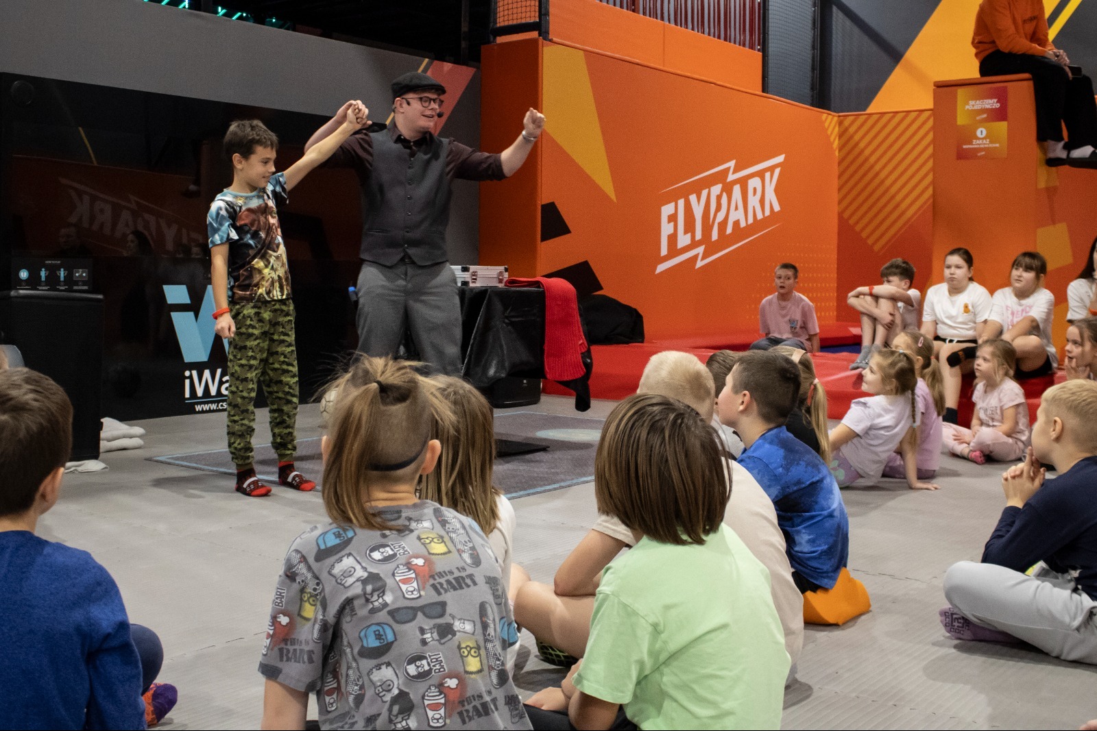 Iluzjonista w kapeluszu prezentuje trik chłopcu na tle logo FlyPark, otoczony grupą dzieci siedzących na podłodze. Scena z imprezy dla dzieci.