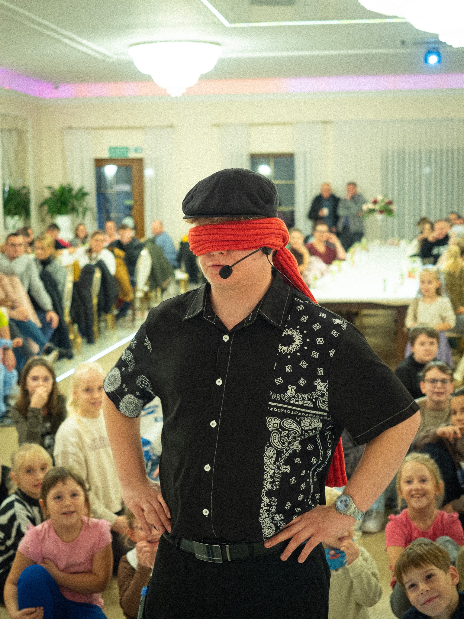 Iluzjonista z zasłoniętymi oczami, w czarnej koszuli, prezentuje się na tle publiczności w Tworogu. Dzieci siedzą na podłodze, dorośli za stołami.