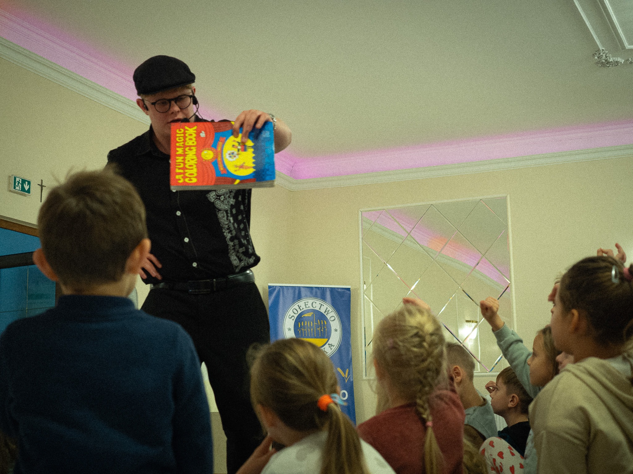 Iluzjonista w czarnym stroju prezentuje kolorowankę magiczną dzieciom. W tle widoczny baner z logo. Sala oświetlona delikatnym, różowym światłem.