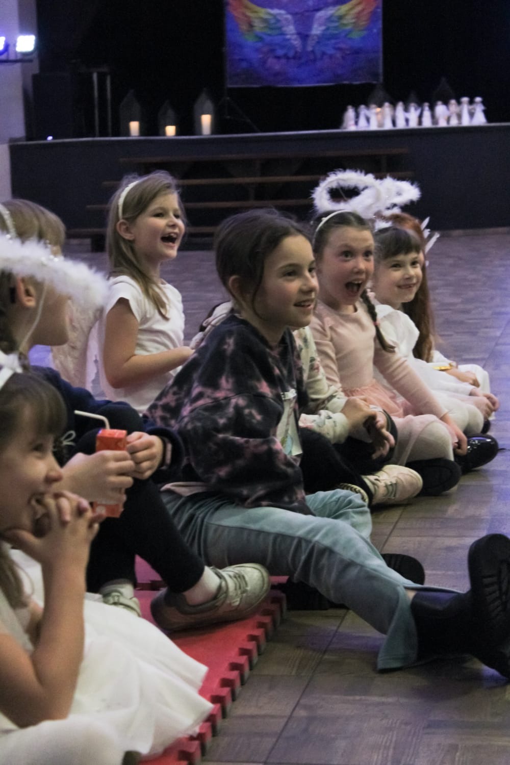 Grupa roześmianych dziewczynek w kostiumach aniołów siedzi rzędem na podłodze podczas wydarzenia. W tle dekoracje sceniczne i oświetlenie.