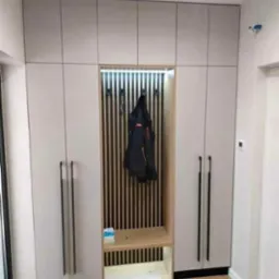 Jasna szafa wnękowa z siedziskiem i oświetleniem LED, czarne, długie uchwyty, drewniane lamele na ścianie wnęki, parkiet na podłodze, kapcie przy szafie.