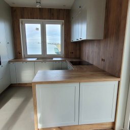 BUDILLO DANIEL KOZIEŁ - Jasna kuchnia z drewnianymi akcentami i blatem, widok na okno z zabudową. Uwagę przyciągają minimalistyczne szafki i ciepłe odcienie drewna.