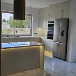 Nowoczesna kuchnia z wyspą i złotym okapem na tle budowy. Jasne szafki z podświetleniem LED i marmurowa podłoga z połyskiem, widok na plac budowy za oknem.