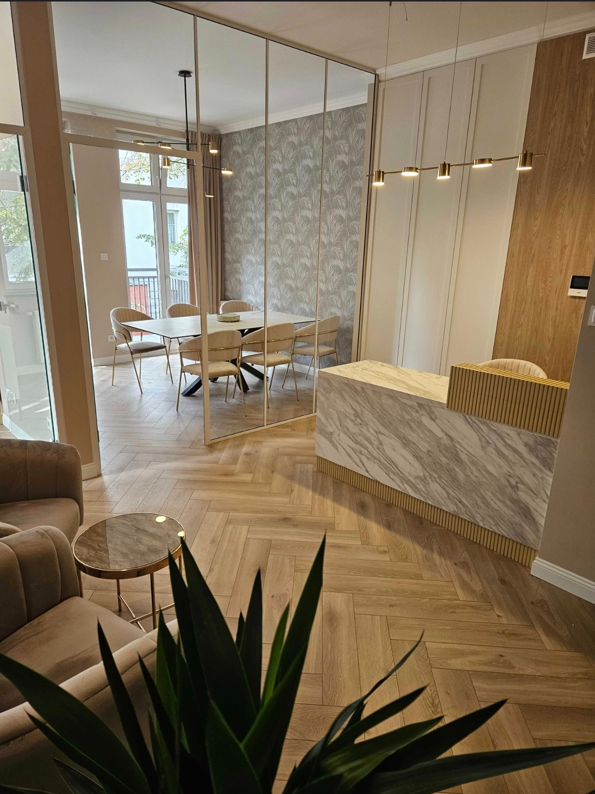 Nowoczesne wykończenie wnętrza: parkiet w jodełkę, marmurowa lada, szklana ściana oddzielająca jadalnię. W tle widoczny balkon i jasne, naturalne światło.