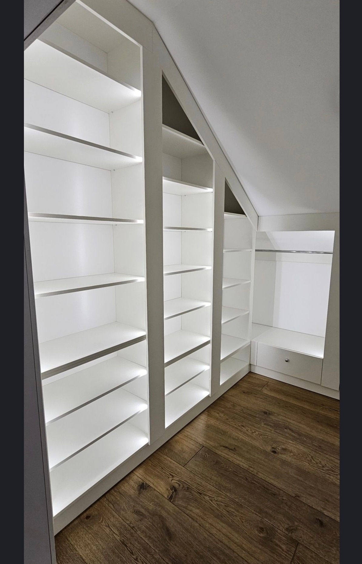 Biała szafa wnękowa na poddaszu z oświetleniem LED, drewniana podłoga. Widok z dołu ukazuje precyzyjne dopasowanie do skosu dachu i eleganckie wykończenie.