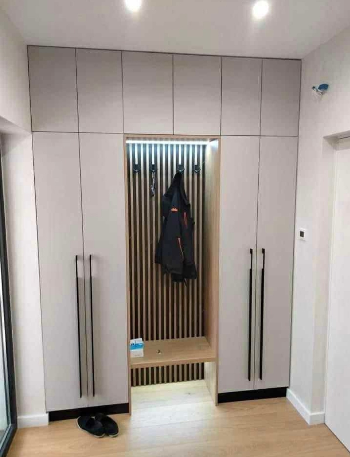 Jasna szafa wnękowa z siedziskiem i oświetleniem LED, czarne, długie uchwyty, drewniane lamele na ścianie wnęki, parkiet na podłodze, kapcie przy szafie.