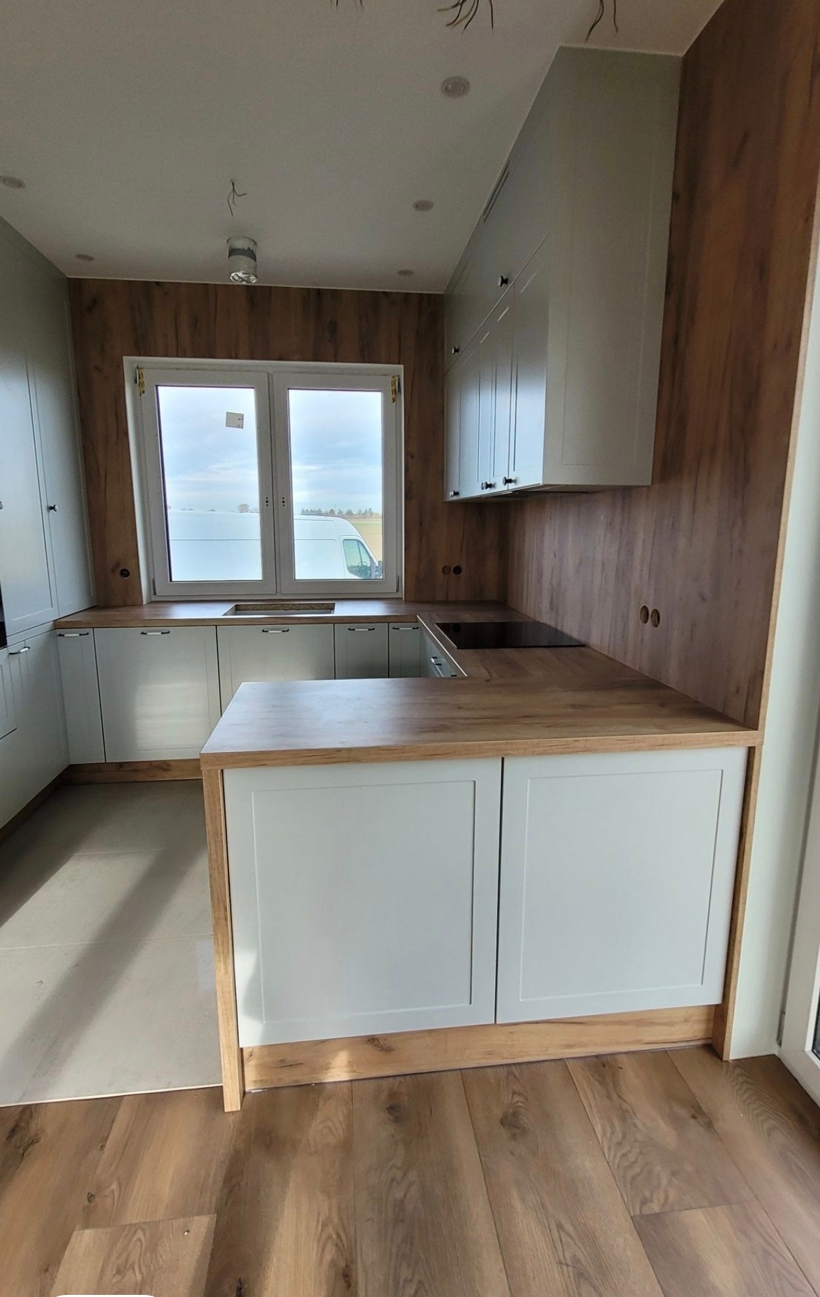 Jasna kuchnia z drewnianymi akcentami i blatem, widok na okno z zabudową. Uwagę przyciągają minimalistyczne szafki i ciepłe odcienie drewna.