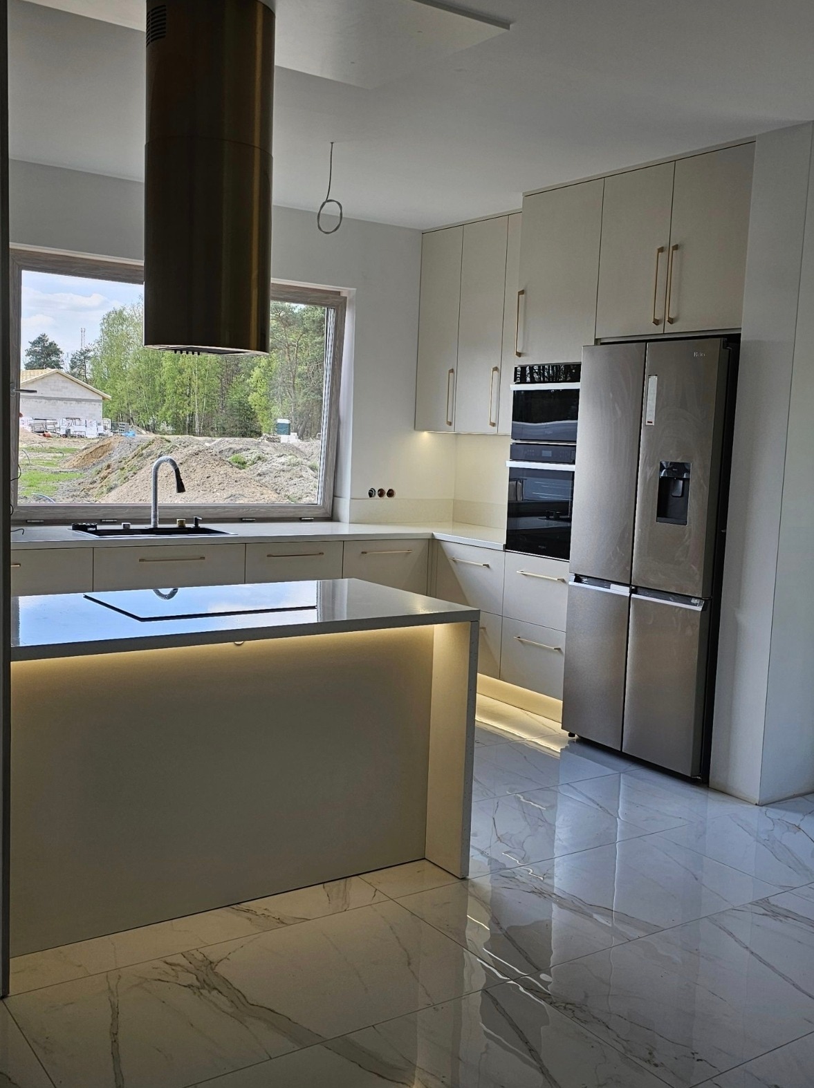 Nowoczesna kuchnia z wyspą i złotym okapem na tle budowy. Jasne szafki z podświetleniem LED i marmurowa podłoga z połyskiem, widok na plac budowy za oknem.