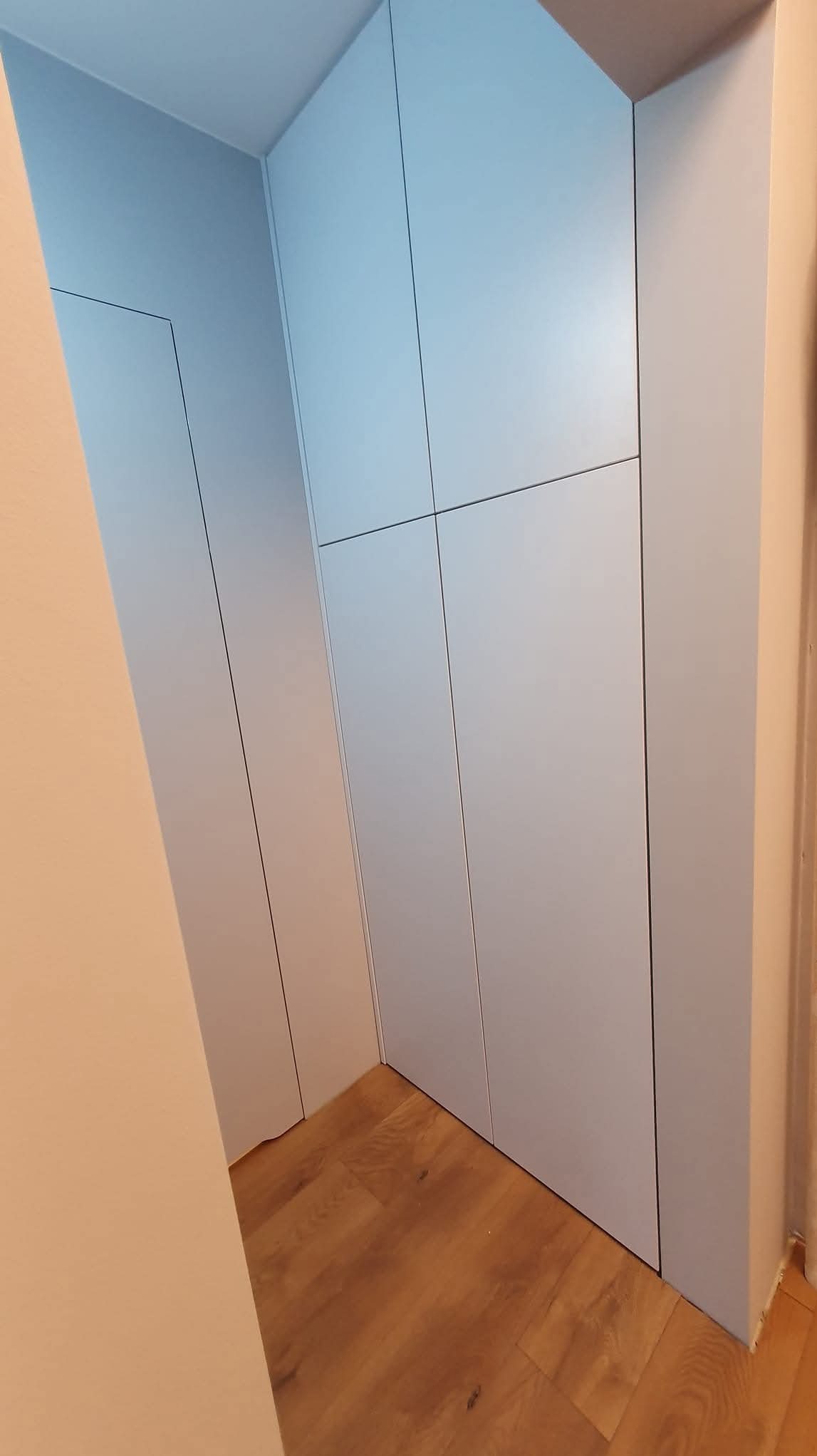 Jasnoszara szafa wnękowa z minimalistycznymi drzwiami, dopasowana do skosu pomieszczenia, na tle drewnianej podłogi. Ujęcie z dołu, podkreśla wysokość zabudowy.