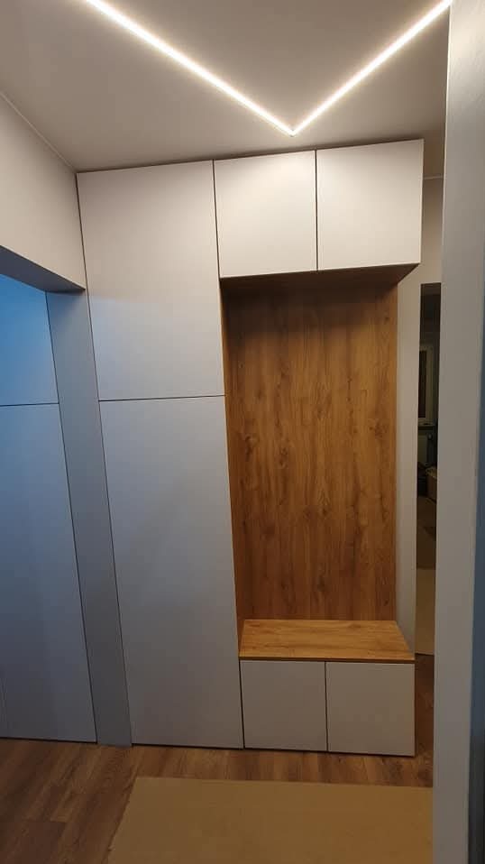 Szara szafa wnękowa z drewnianym panelem i siedziskiem, oświetlona ledami, w przedpokoju. Minimalistyczny design, zabudowa pod sufit.