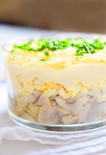 Sałatka śledziowa warstwowa w szklanym naczyniu, z posiekanym śledziem, jajkiem, warzywami i majonezem, udekorowana zieleniną. Jasne tło, delikatne światło.