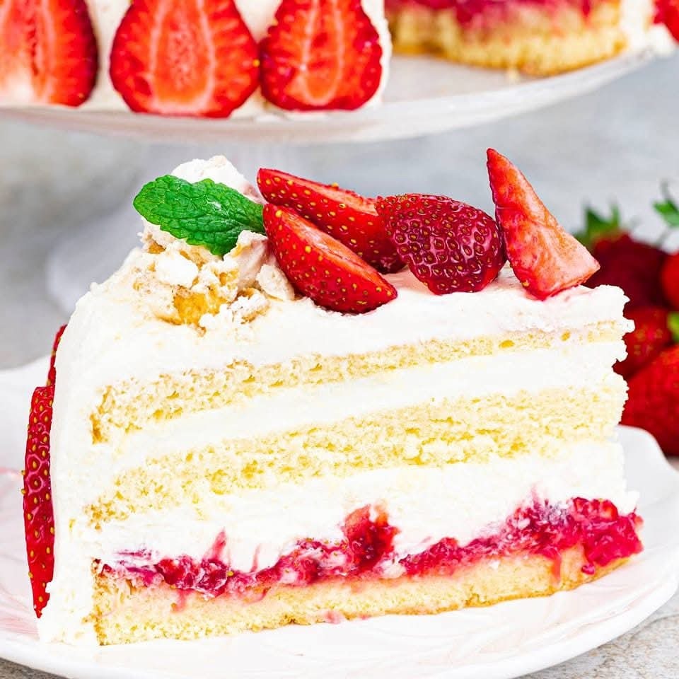 Kremowy tort z truskawkami na białym talerzu, dekorowany miętą. Widoczny przekrój z warstwami biszkoptu, kremu i truskawek. W tle tort na paterze.