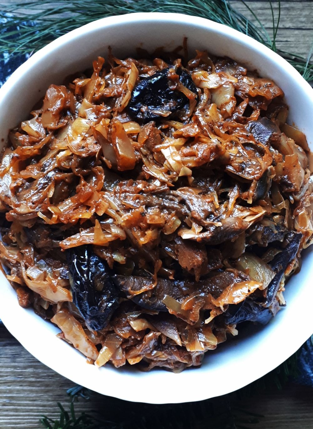 Wigilijna kapusta z grzybami i śliwkami w białej misce, udekorowana gałązkami świerku. Potrawa gotowa do spożycia, na drewnianym stole.