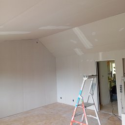 Malermester RAF Wojciechowski - Wnętrze pokoju z zamontowaną płytą kartonowo-gipsową na ścianach i suficie, widoczne łączenia płyt zaszpachlowane, drabina aluminiowa stoi w rogu, widoczne otwory na gniazdka elektryczne w ścianie.