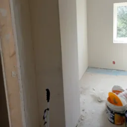 Pokój w trakcie wykańczania, widoczne świeżo położone gładzie na ścianach, podłoga zabezpieczona folią, puszki z farbą i narzędzia malarskie.