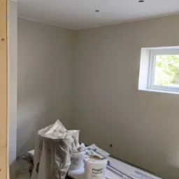 Świeżo pomalowane ściany w odcieniach beżu, widoczne wiaderka z farbą Flügger Flutex 2S, zakryte meble folią malarską i biała listwa przysufitowa.