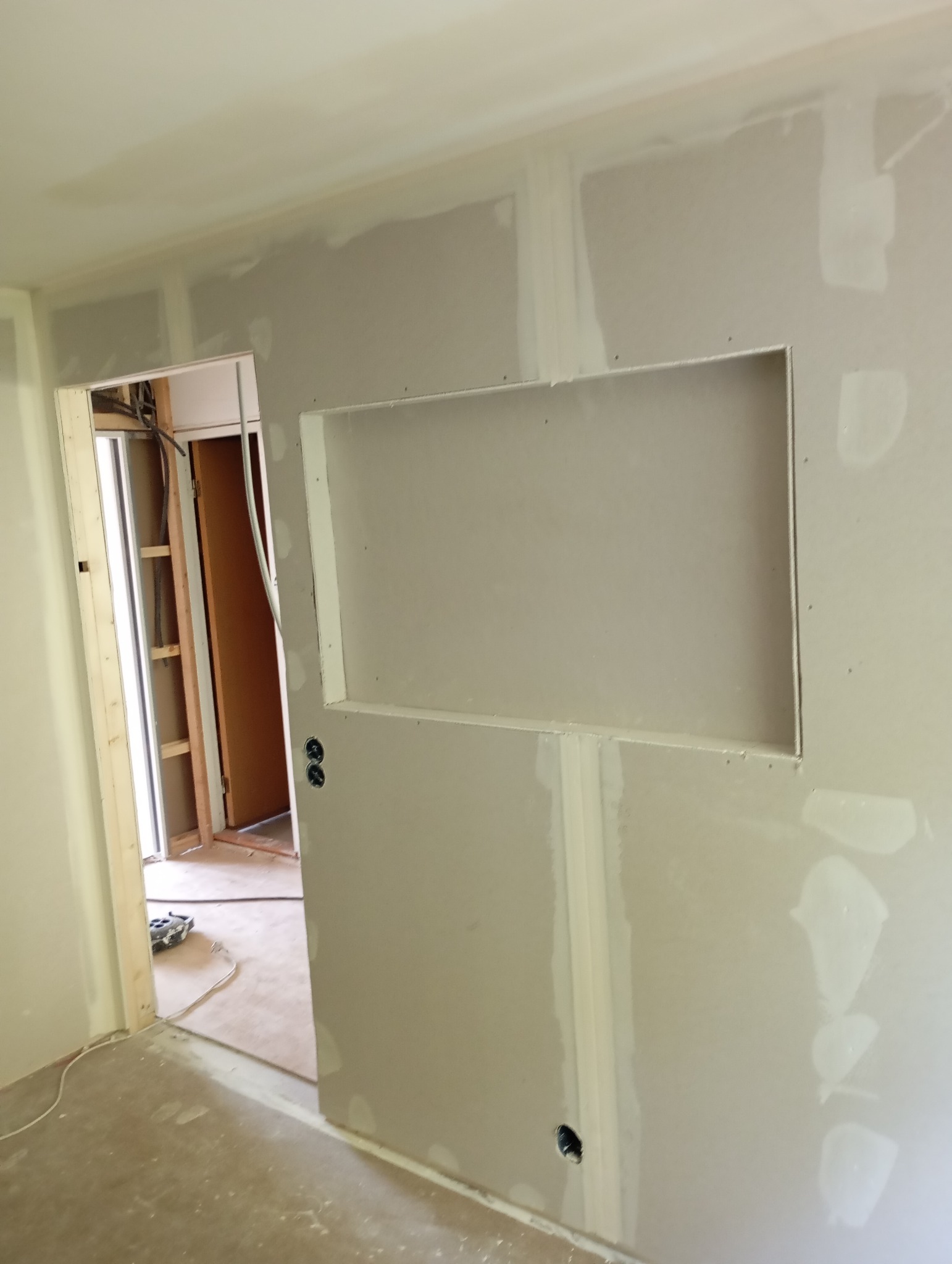 Ściana z płyty kartonowo-gipsowej z wnęką w trakcie szpachlowania, widoczne otwory na gniazdka elektryczne i narzędzia w tle.