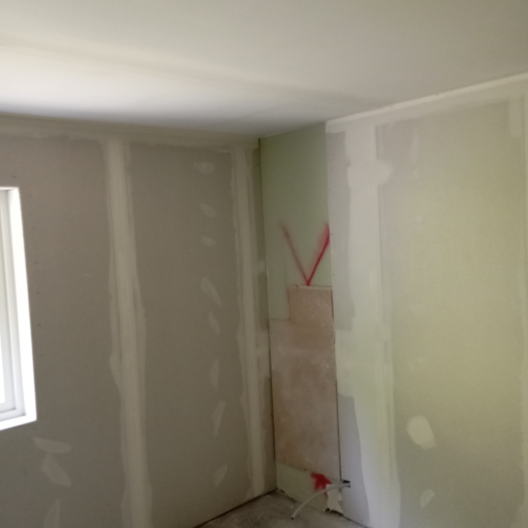 Wnętrze w trakcie montażu płyt karton-gips na ścianach, widoczne łączenia i szpachlowanie, czerwone oznaczenia na ścianie, okno z lewej strony.