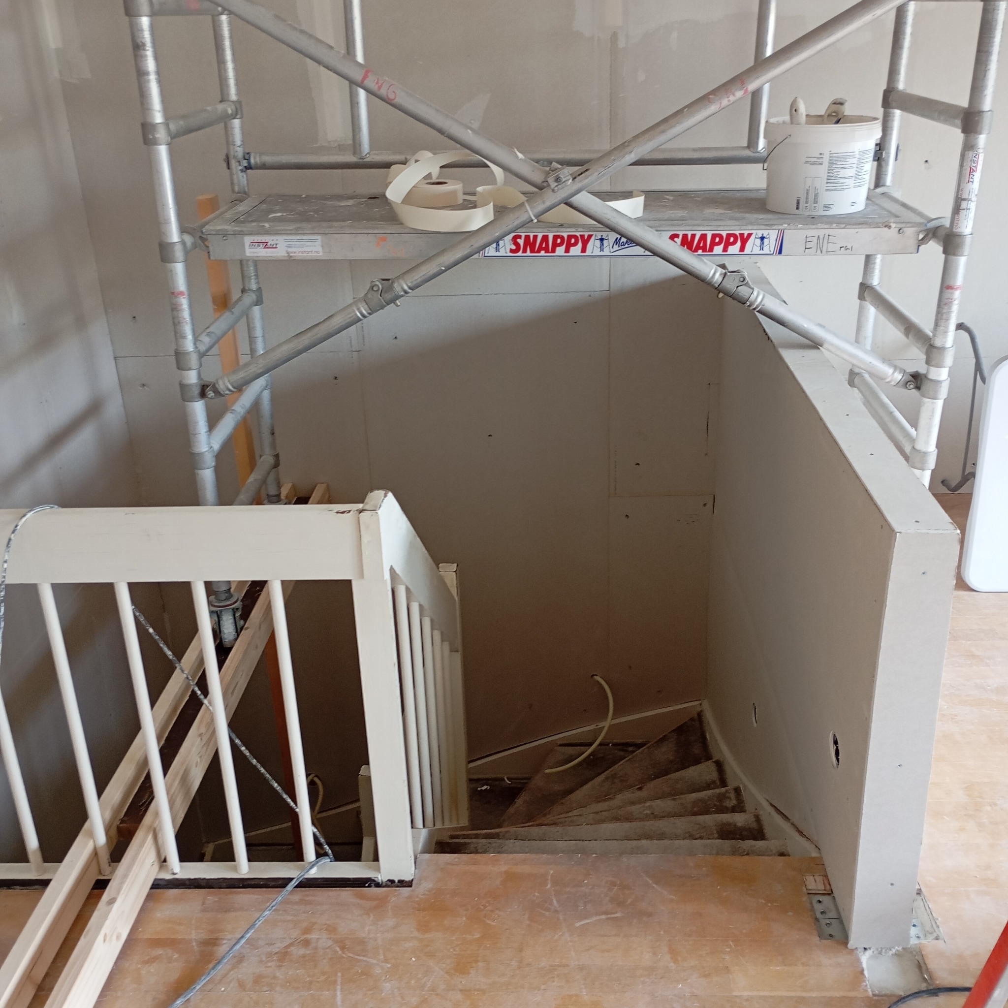 Montaż płyt karton-gips na ścianie przy schodach, widoczne rusztowanie, wiadro z gładzią i rolka taśmy na podeście rusztowania, surowe drewniane schody.