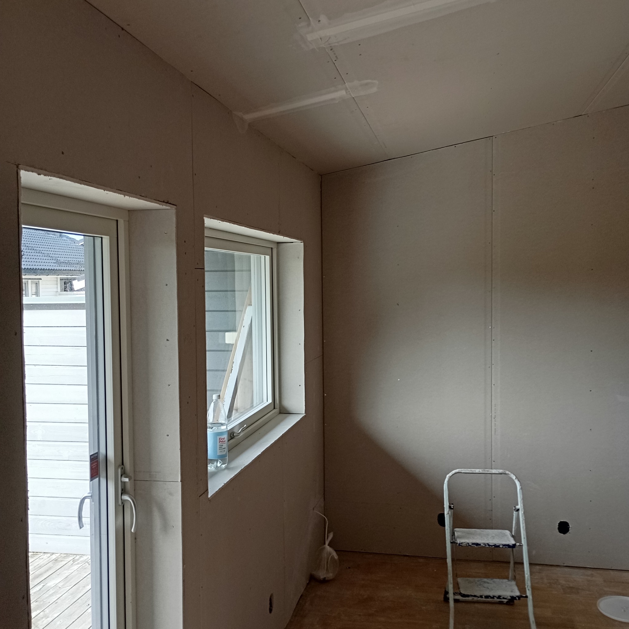 Pokój w trakcie wykańczania, ściany obłożone płytami karton-gips, widoczne łączenia płyt na suficie wzmocnione taśmą, okno z butelką wody na parapecie, drabina malarska oparta o ścianę.