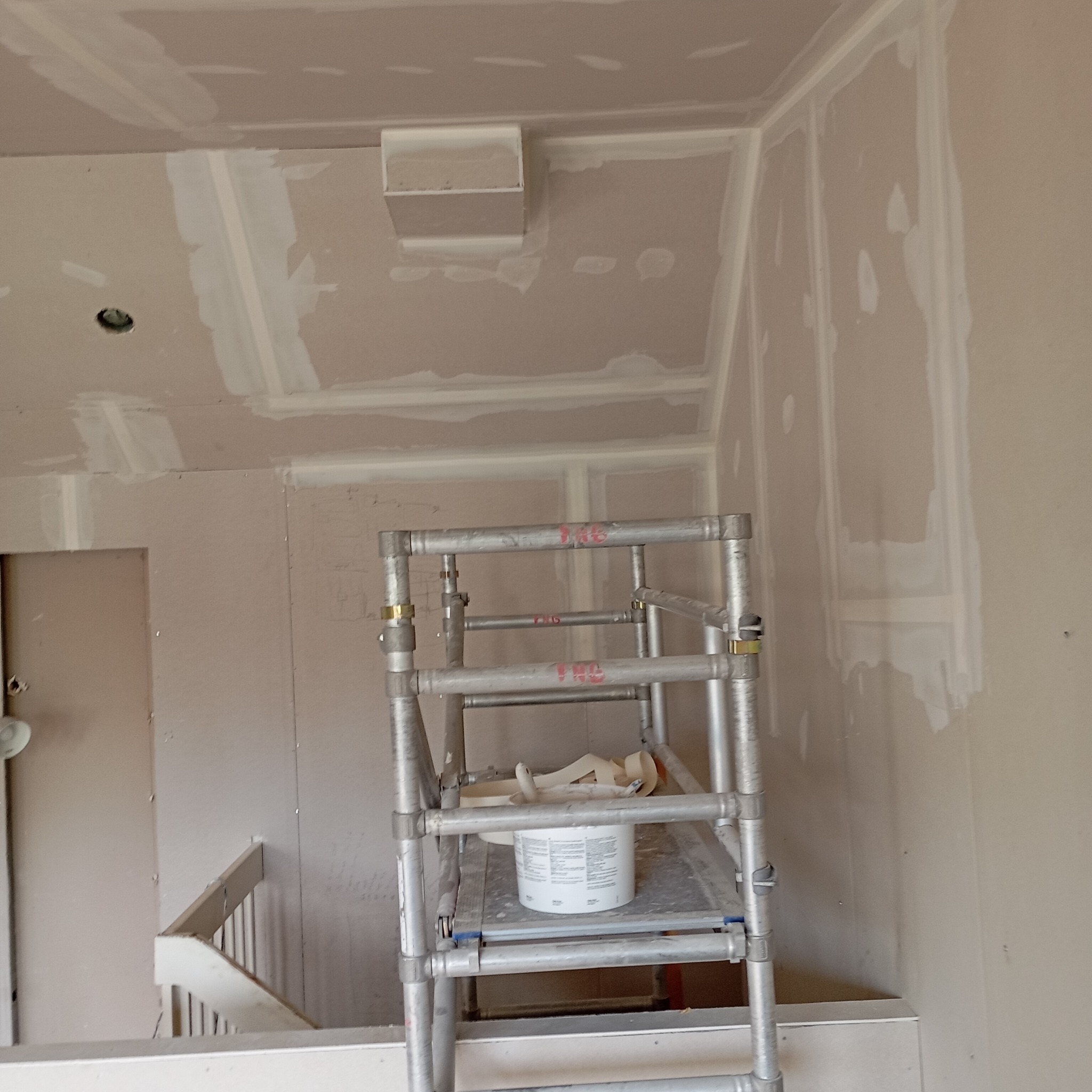 Ściana z płyt kartonowo-gipsowych w trakcie szpachlowania, widoczne łączenia i narożniki zabezpieczone taśmą, metalowy ruszt z wiadrem i narzędziami w tle, fragment schodów w dolnej części kadru.