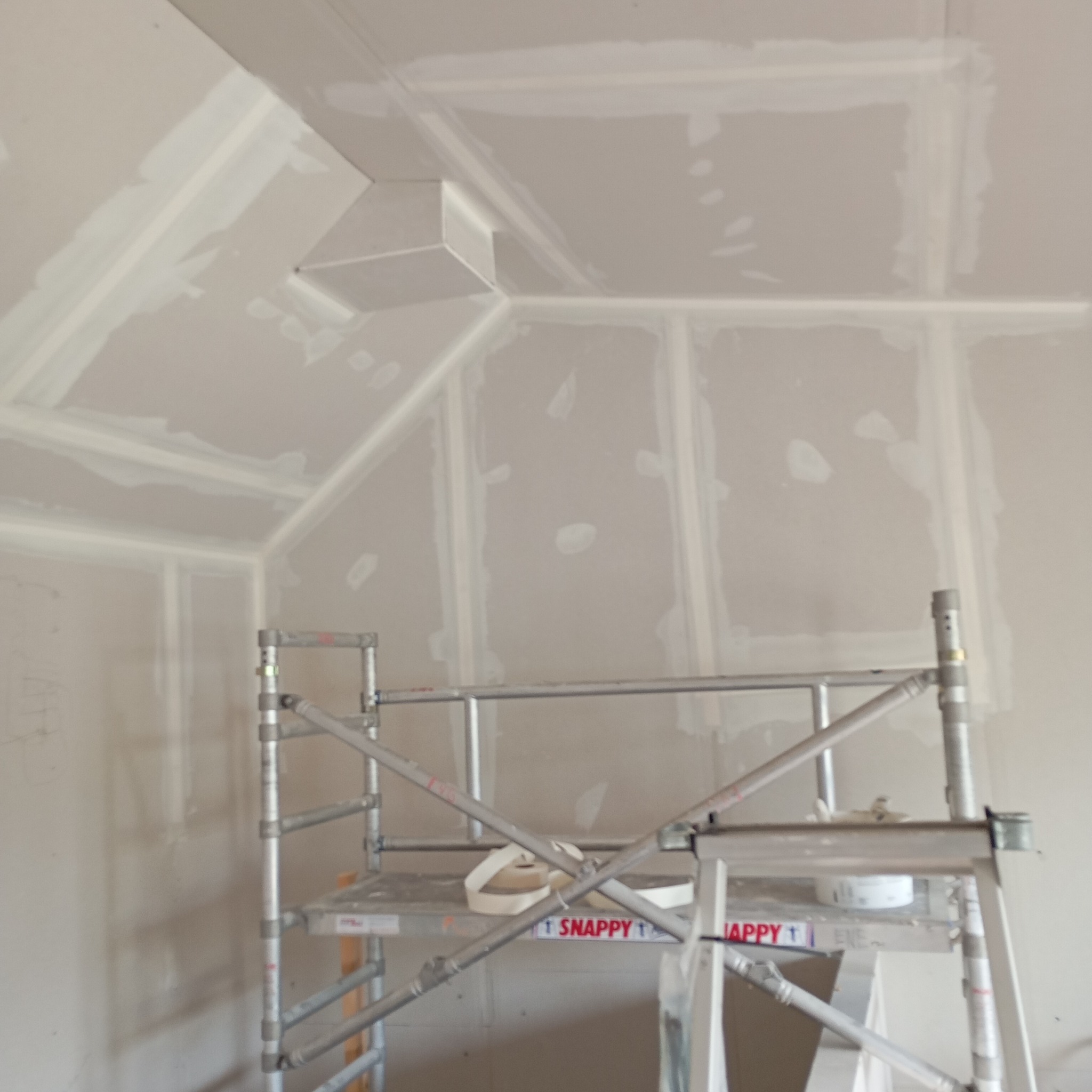 Sufit poddasza z zamontowaną płytą kartonowo-gipsową, z widocznymi łączeniami zaszpachlowanymi i częściowo wyszlifowanymi, ustawione rusztowanie z rolką taśmy do spoinowania.