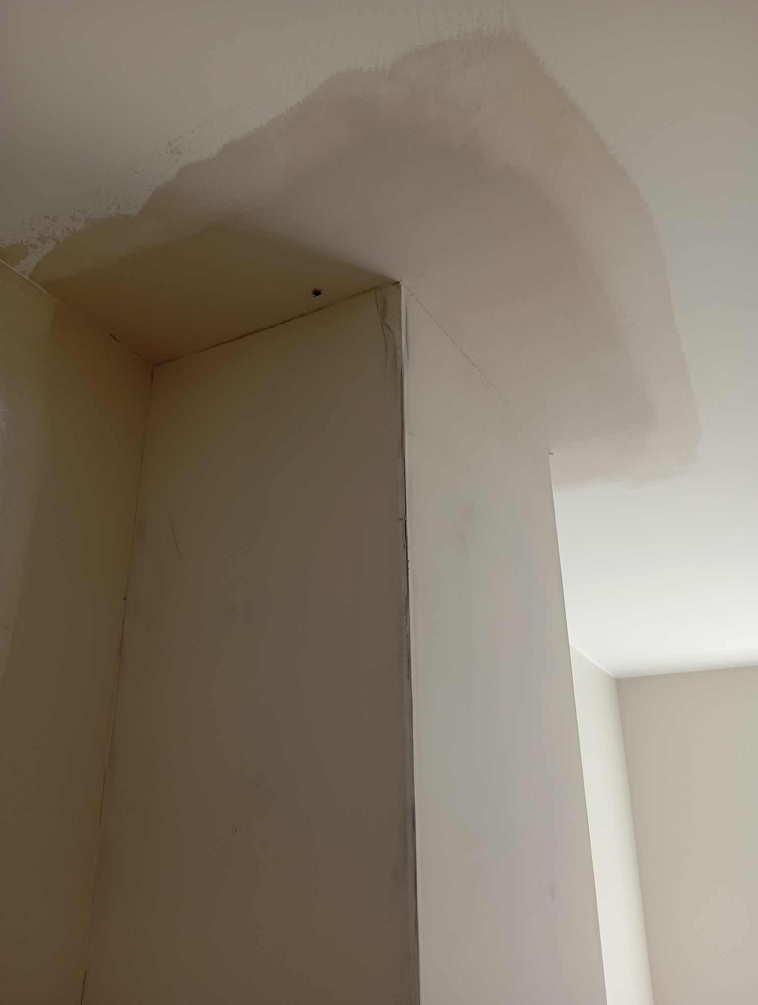 Zacieki na suficie i ścianie w rogu pomieszczenia, widoczne ślady po zalaniu, jasne ściany i sufit, perspektywa z dołu.