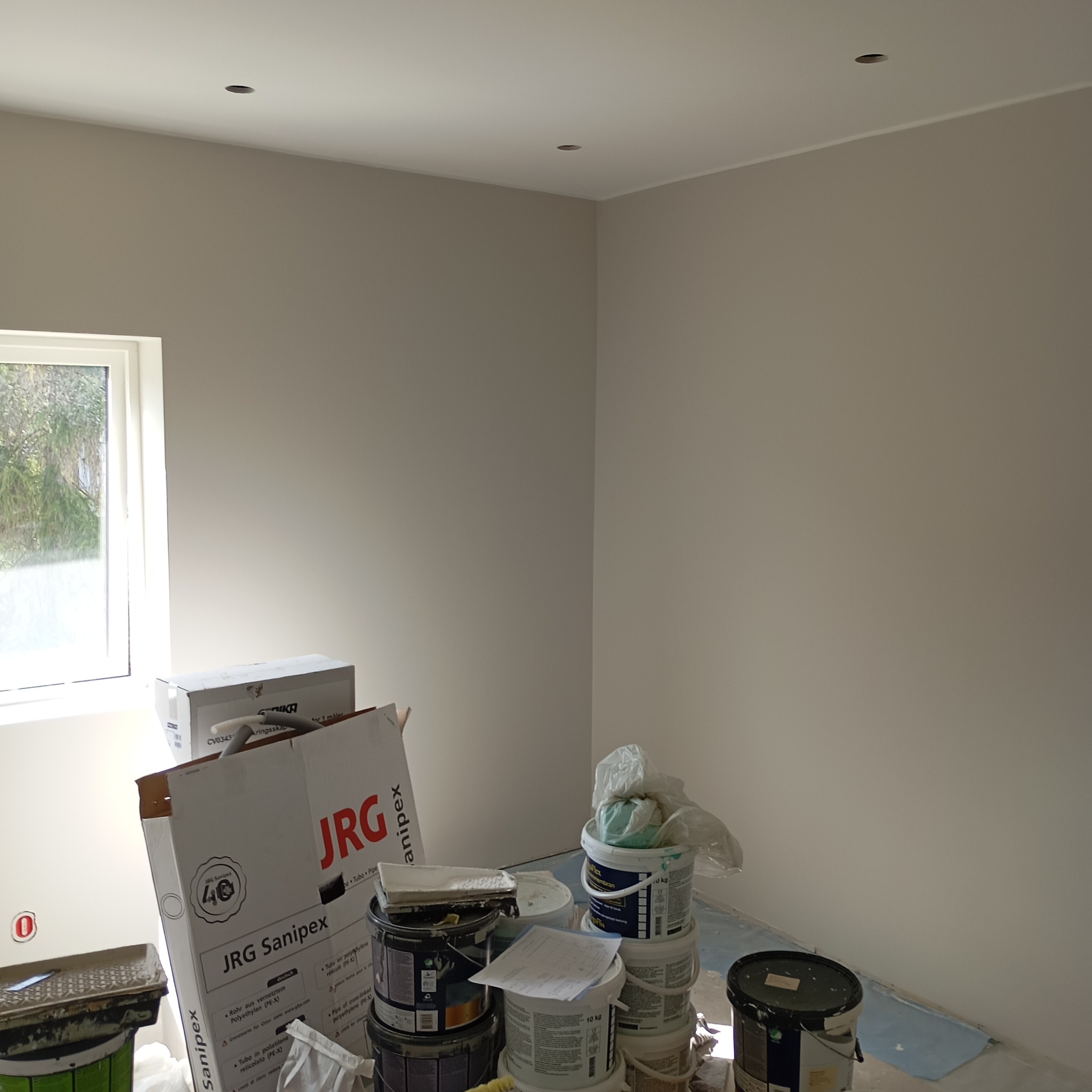 Pokój po malowaniu ścian na jasnoszaro, widoczne puszki z farbą, kartony i akcesoria malarskie, przygotowane do sprzątania po remoncie.