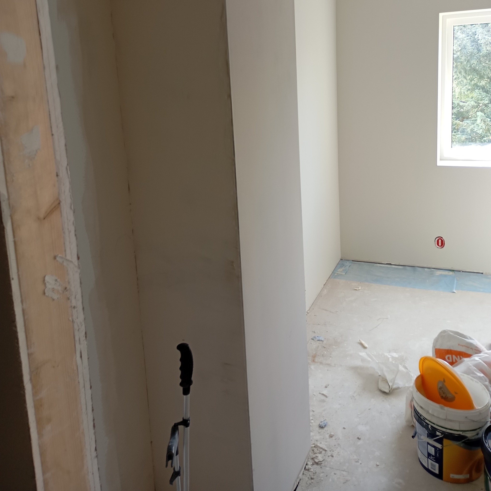 Pokój w trakcie wykańczania, widoczne świeżo położone gładzie na ścianach, podłoga zabezpieczona folią, puszki z farbą i narzędzia malarskie.