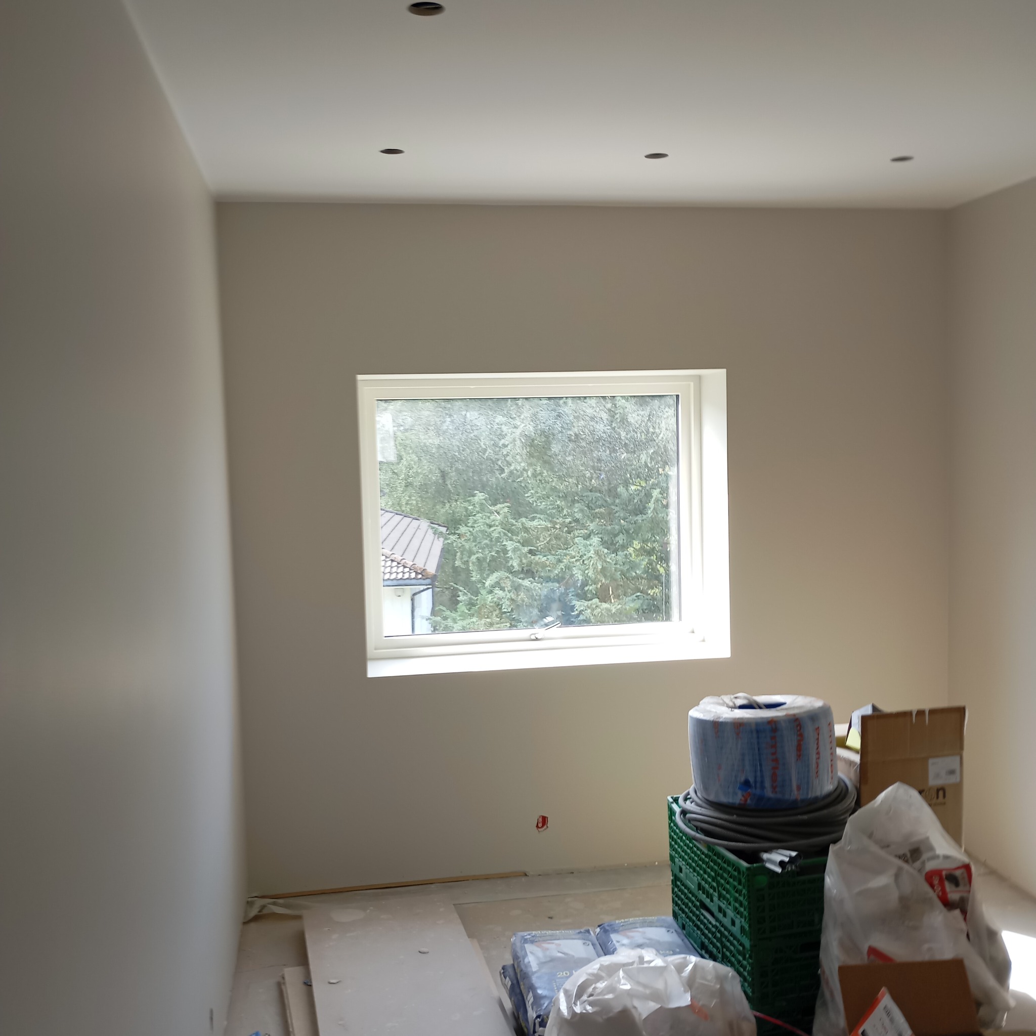 Pokój w trakcie wykańczania, widoczne gładkie, pomalowane ściany, okno z widokiem na zieleń, materiały budowlane w workach i rolkach, puszka elektryczna wystająca ze ściany.
