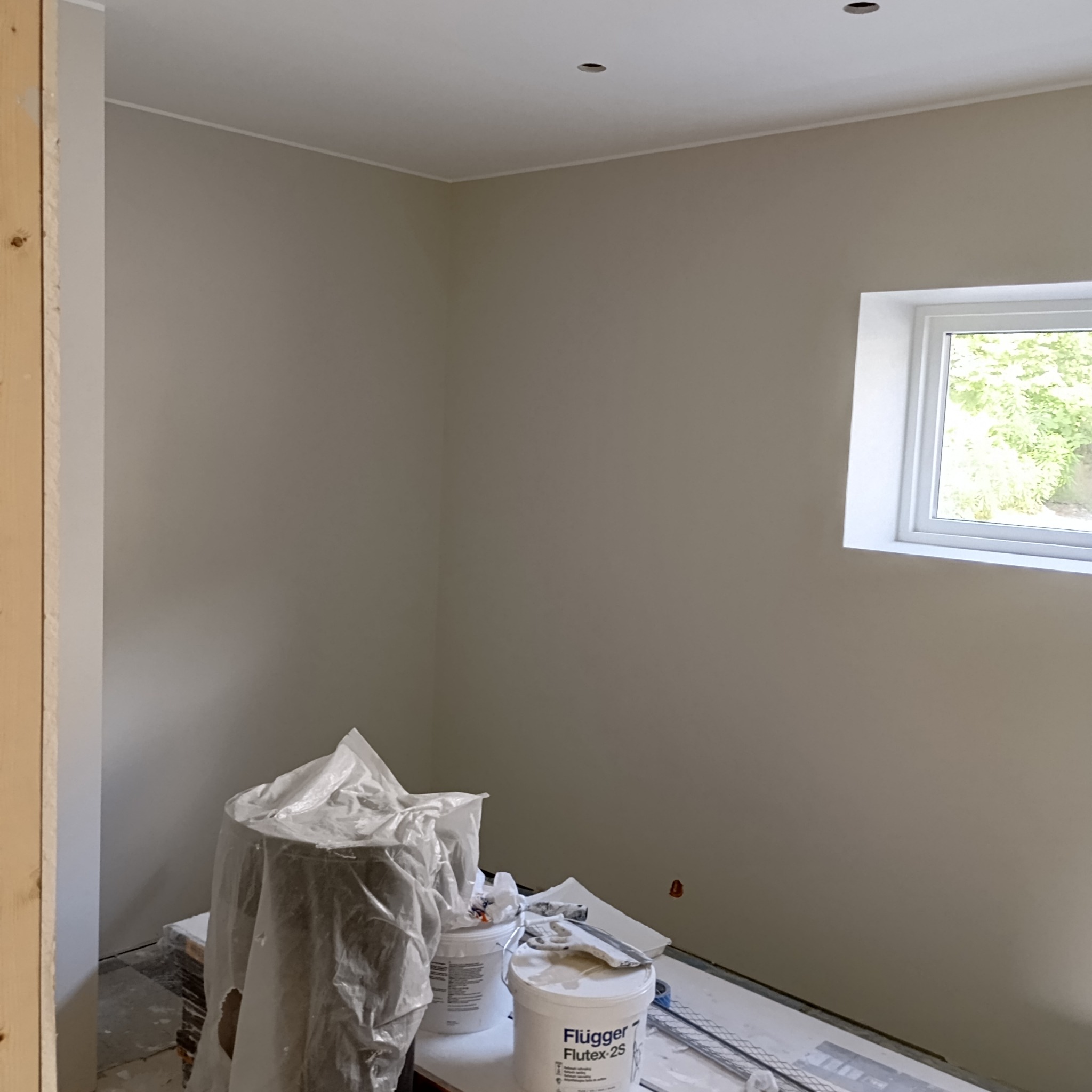 Świeżo pomalowane ściany w odcieniach beżu, widoczne wiaderka z farbą Flügger Flutex 2S, zakryte meble folią malarską i biała listwa przysufitowa.